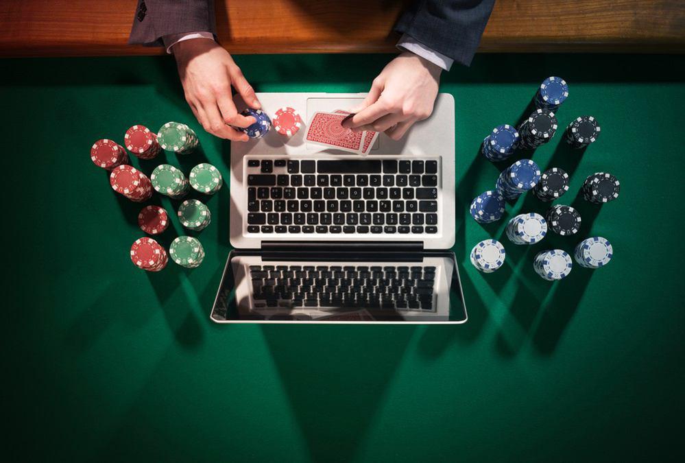 A top-down view of a poker table with a laptop in the center. A person in a suit is holding a playing card and a poker chip over the laptop keyboard.